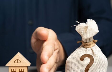 Hand in between house model and a bag of money with question mark printed on it