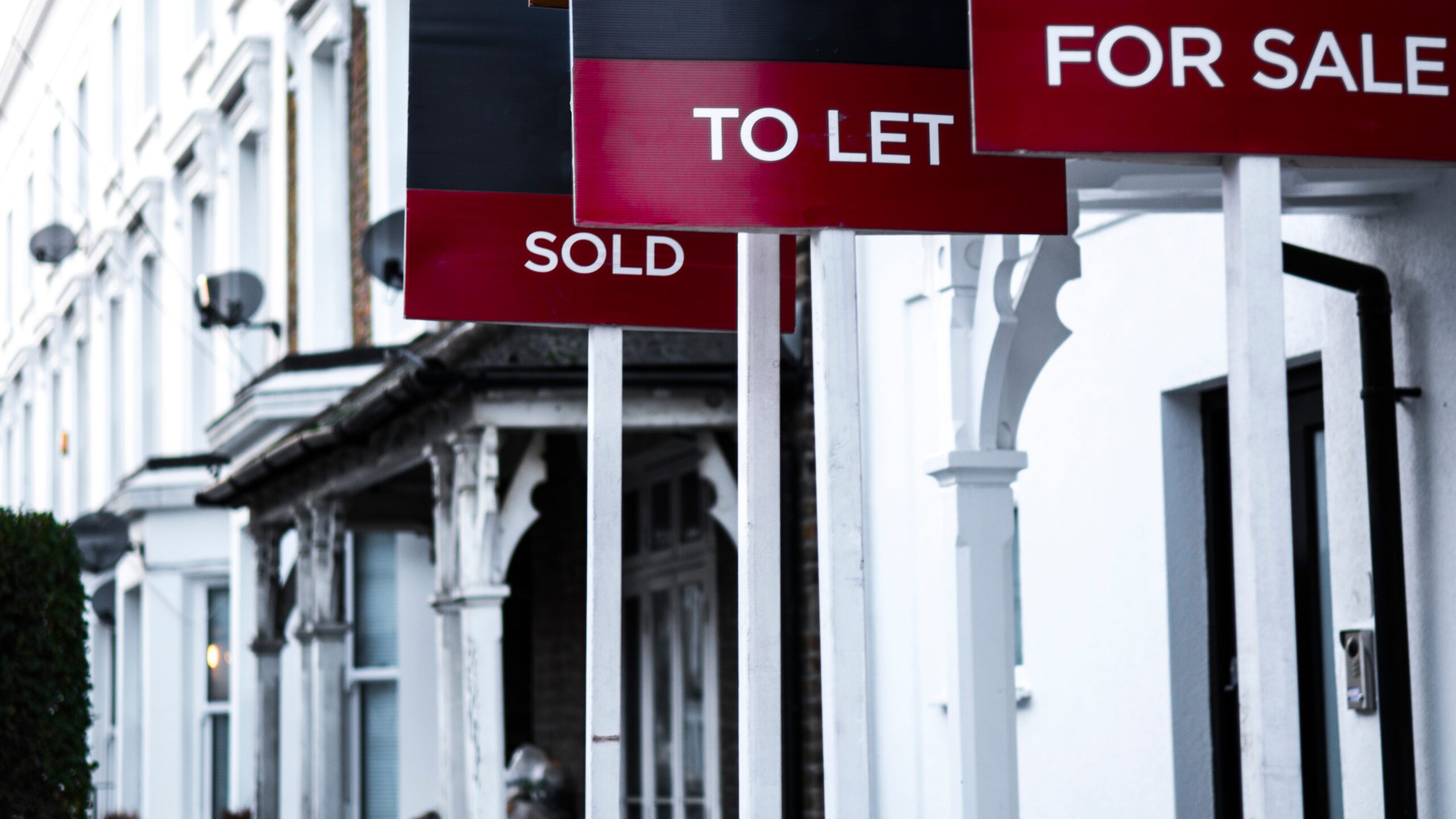 A row of real estate signs in front of a row of houses. The signs read "To Let," "For Sale," and "Sold."