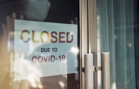 Business office or store shop is closed, bankrupt business due to the effect of novel Coronavirus (COVID-19) pandemic. Unidentified person wearing mask hanging closed sign in background on front door.