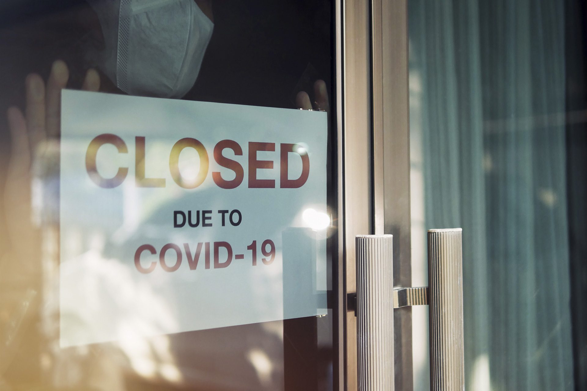 Business office or store shop is closed, bankrupt business due to the effect of novel Coronavirus (COVID-19) pandemic. Unidentified person wearing mask hanging closed sign in background on front door.