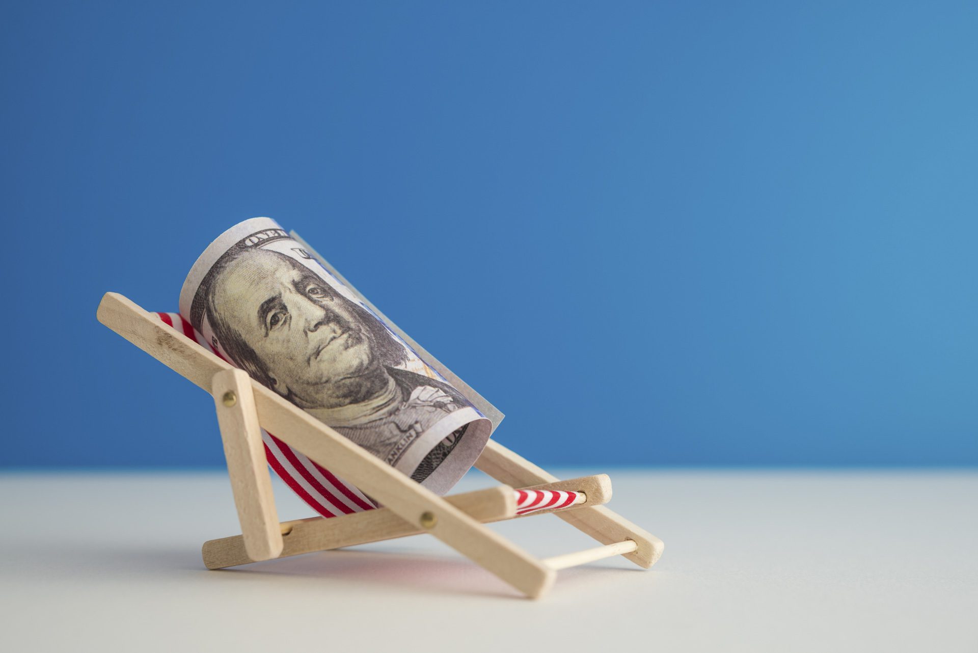 Happy US dollar bill banknote rest on wooden beach chair with blue background copy space in summer beach holiday. Financial freedom through passive income, success wealth from investment concept.