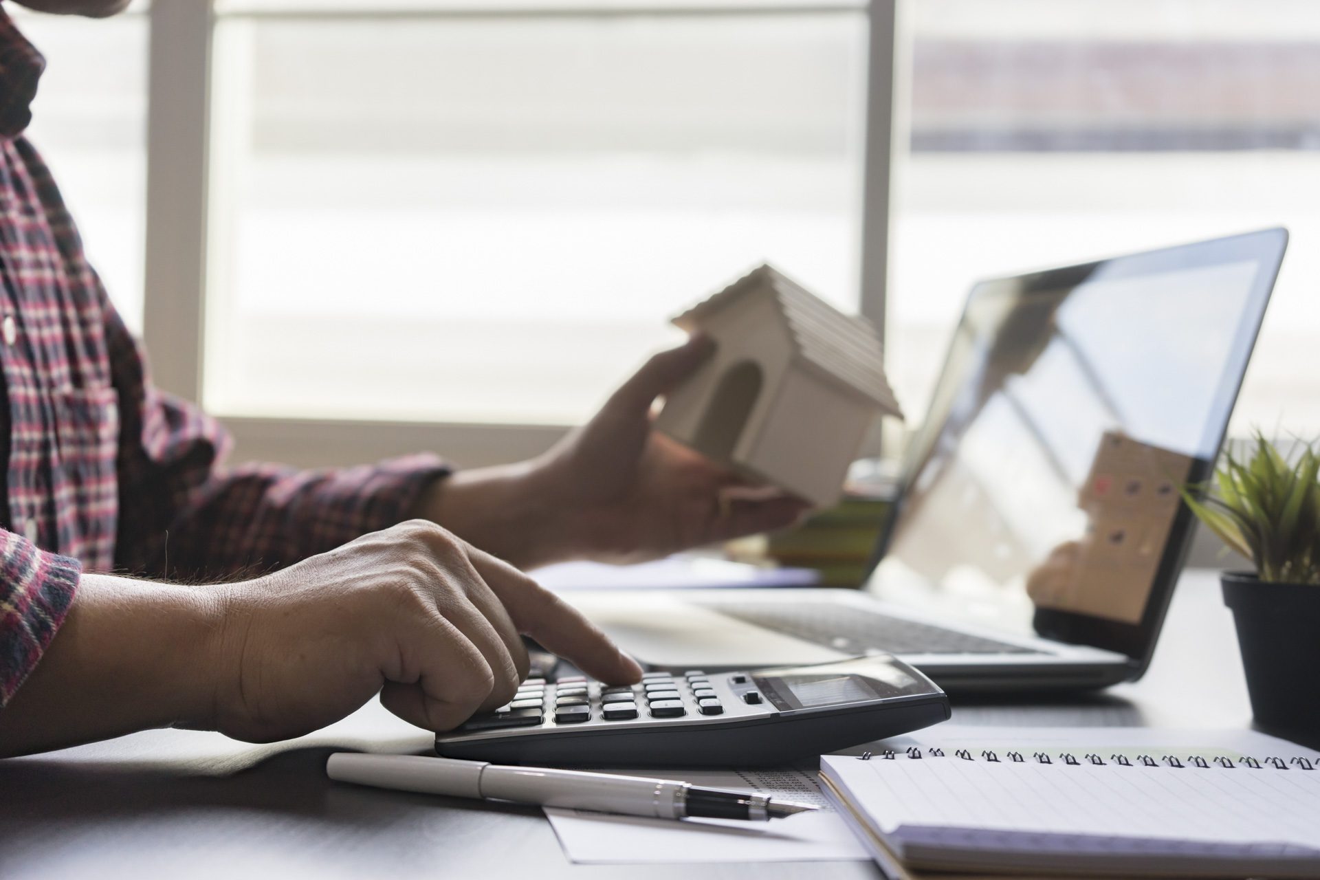 Property investor holding wooden house and doing calculations while researching the real estate market on laptop
