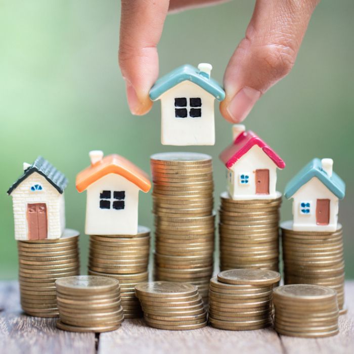 A hand places a miniature house on top of a stack of coins. Behind it are several other stacks of coins each topped with a miniature house.